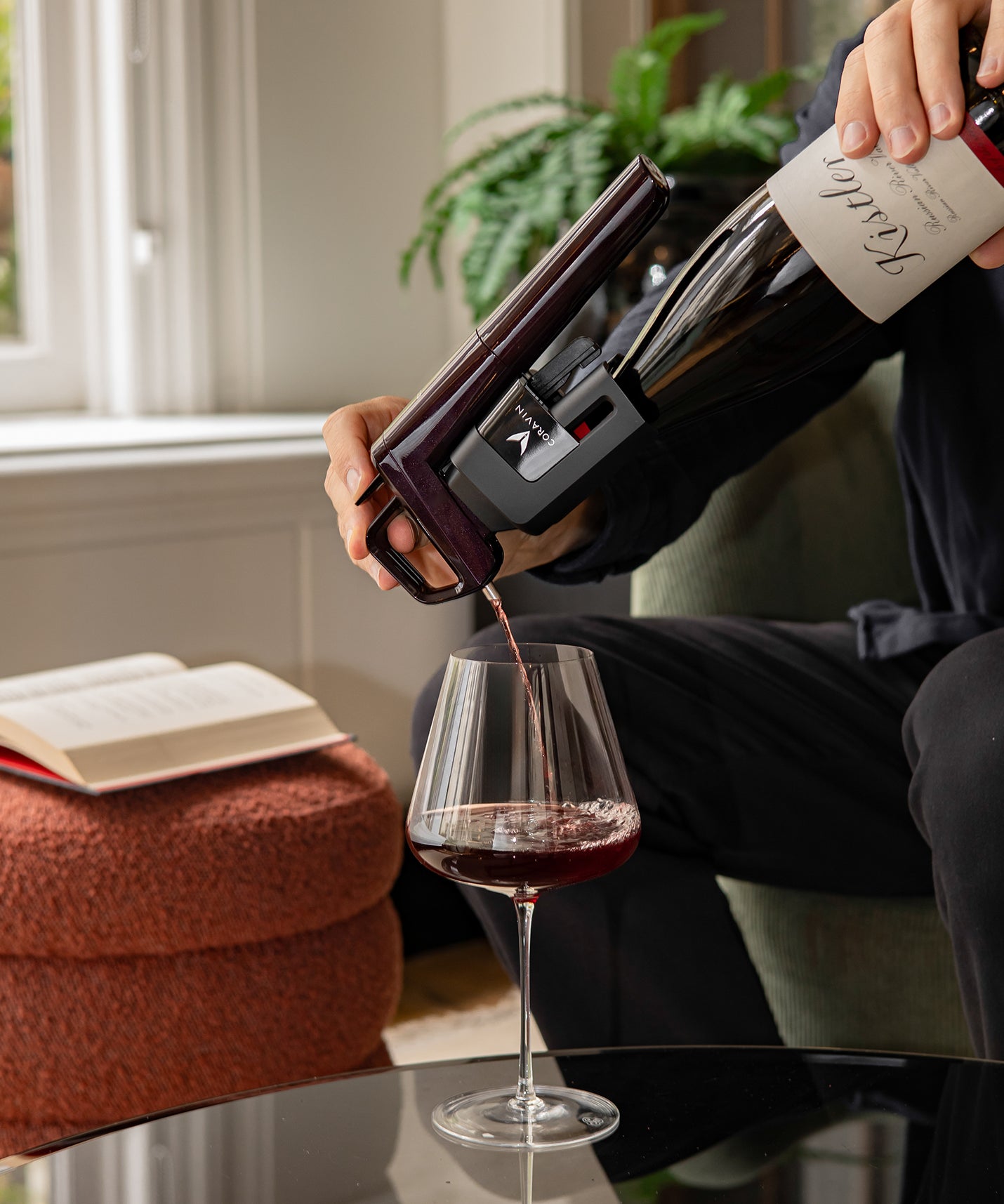 Person pouring red wine from a bottle into a glass, with a book and plant in the background.