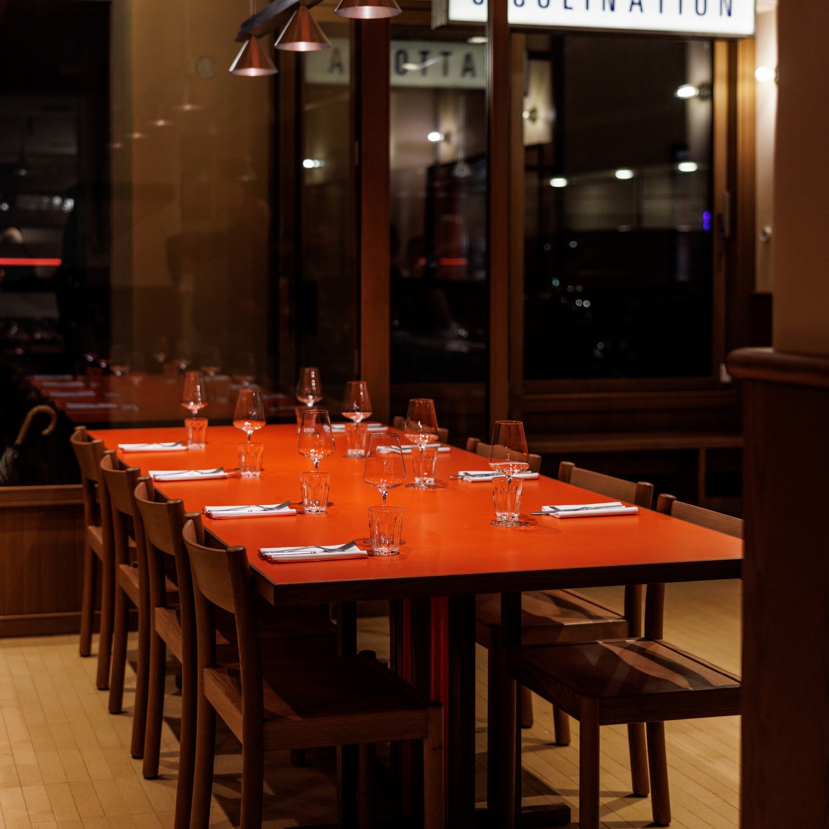 A brightly lit restaurant interior features a large orange table set for dinner with glasses and utensils, under hanging lights.